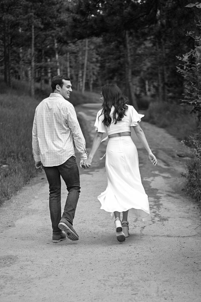 Black and white image of a couple walking hand in hand down a forest path, glancing back with smiles.