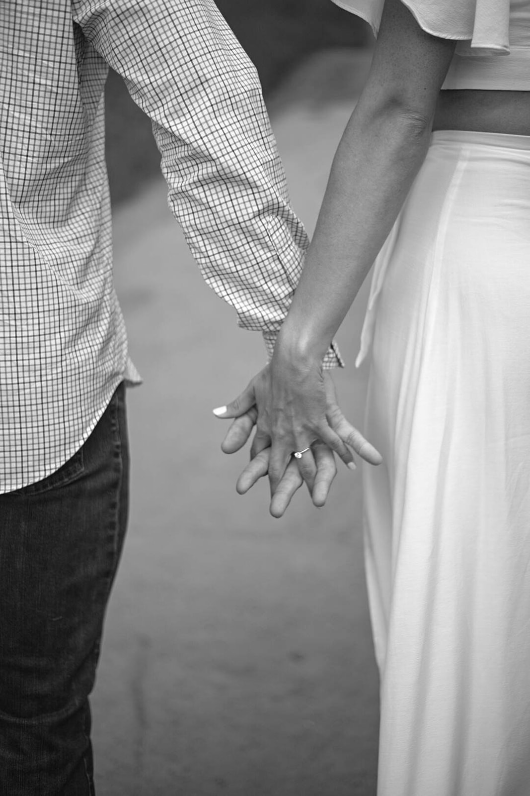 Black-and-white image of a couple walking hand in hand down a trail, fingers interlaced.