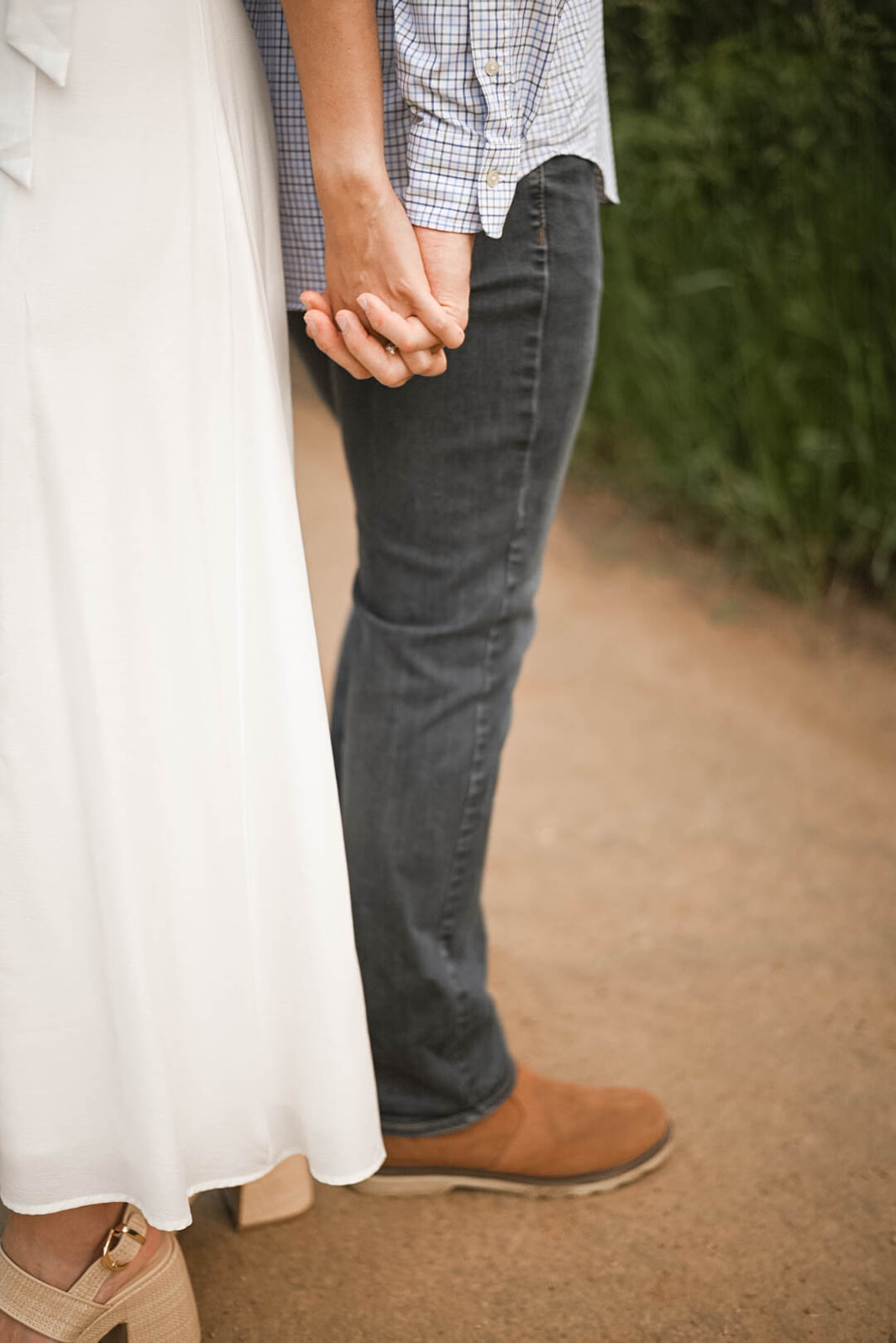 Close-up of couple’s hands interlocked, showing her engagement ring while standing on a dirt path.