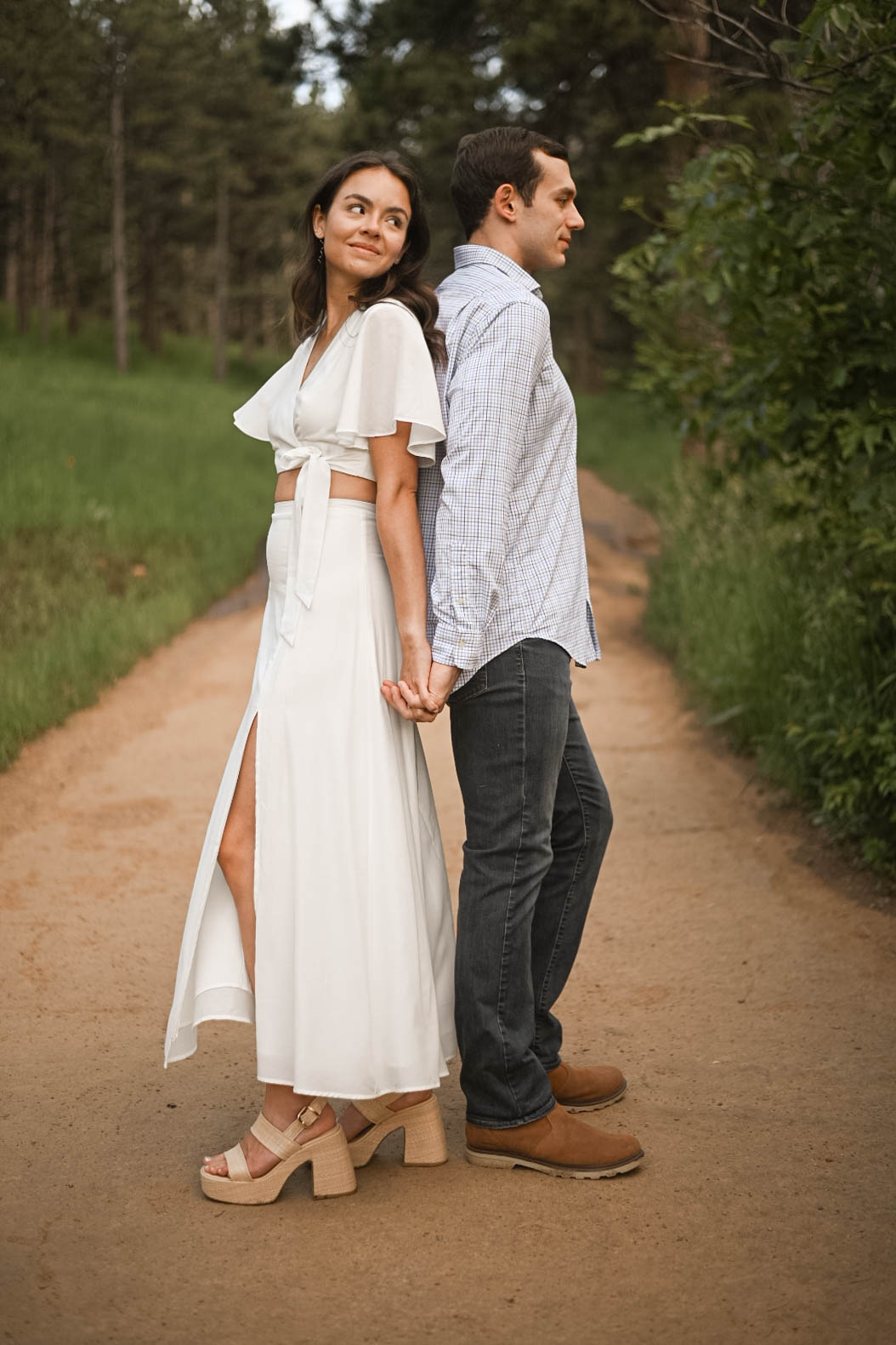 Couple standing back-to-back on a dirt path, holding hands, with trees surrounding them.