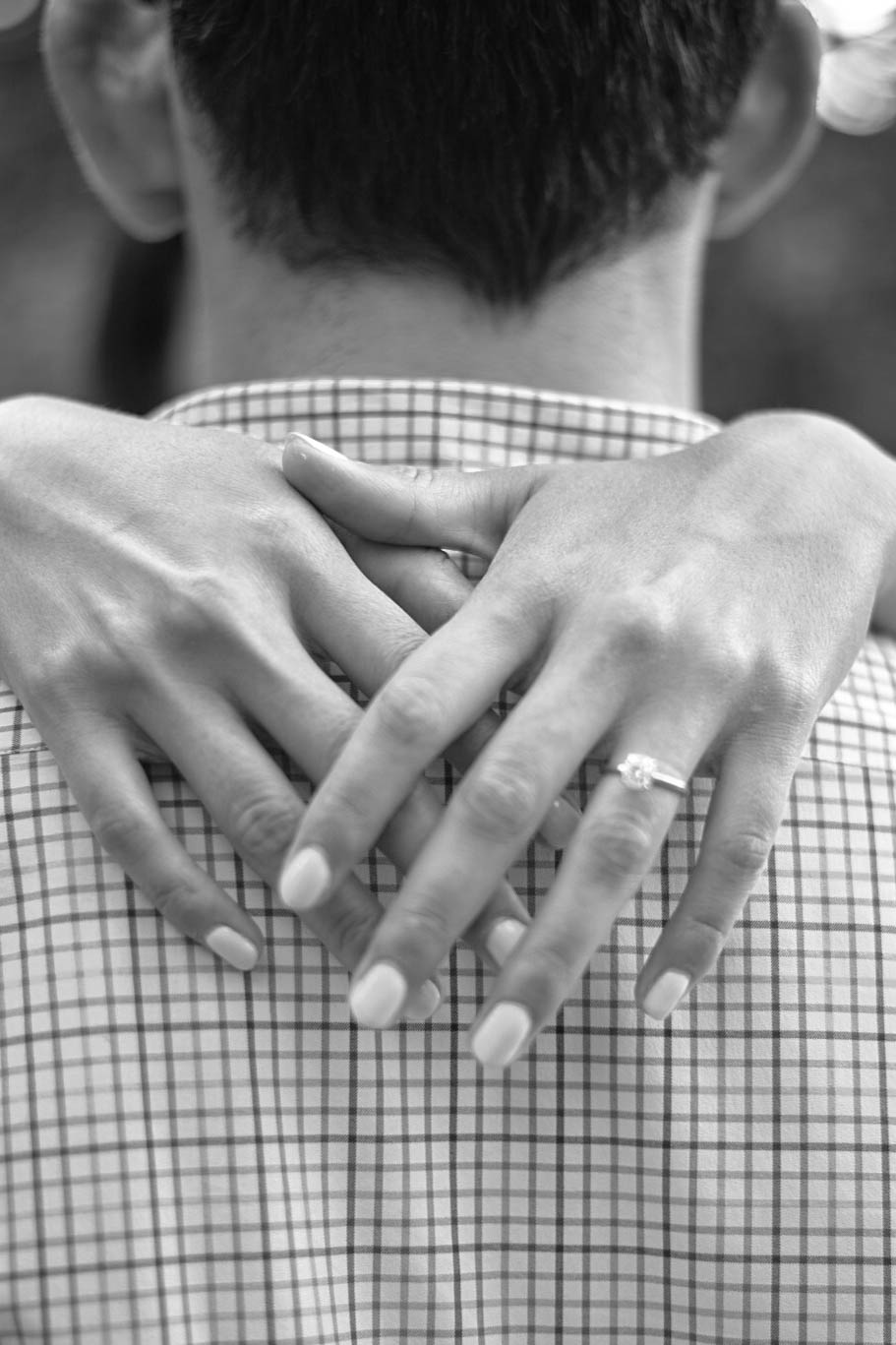 Black-and-white photo of a woman’s hands with a diamond engagement ring resting on her partner’s back.