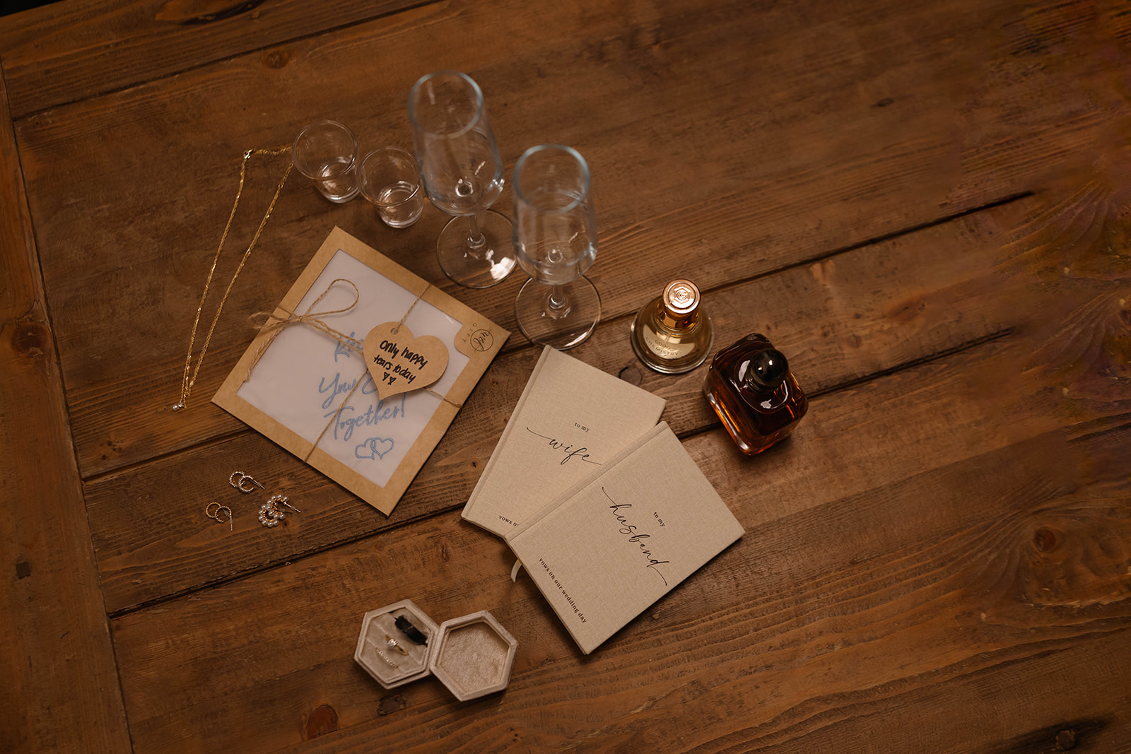 Flat lay of wedding details on a rustic wooden table, including vow books, rings, jewelry, and champagne glasses.