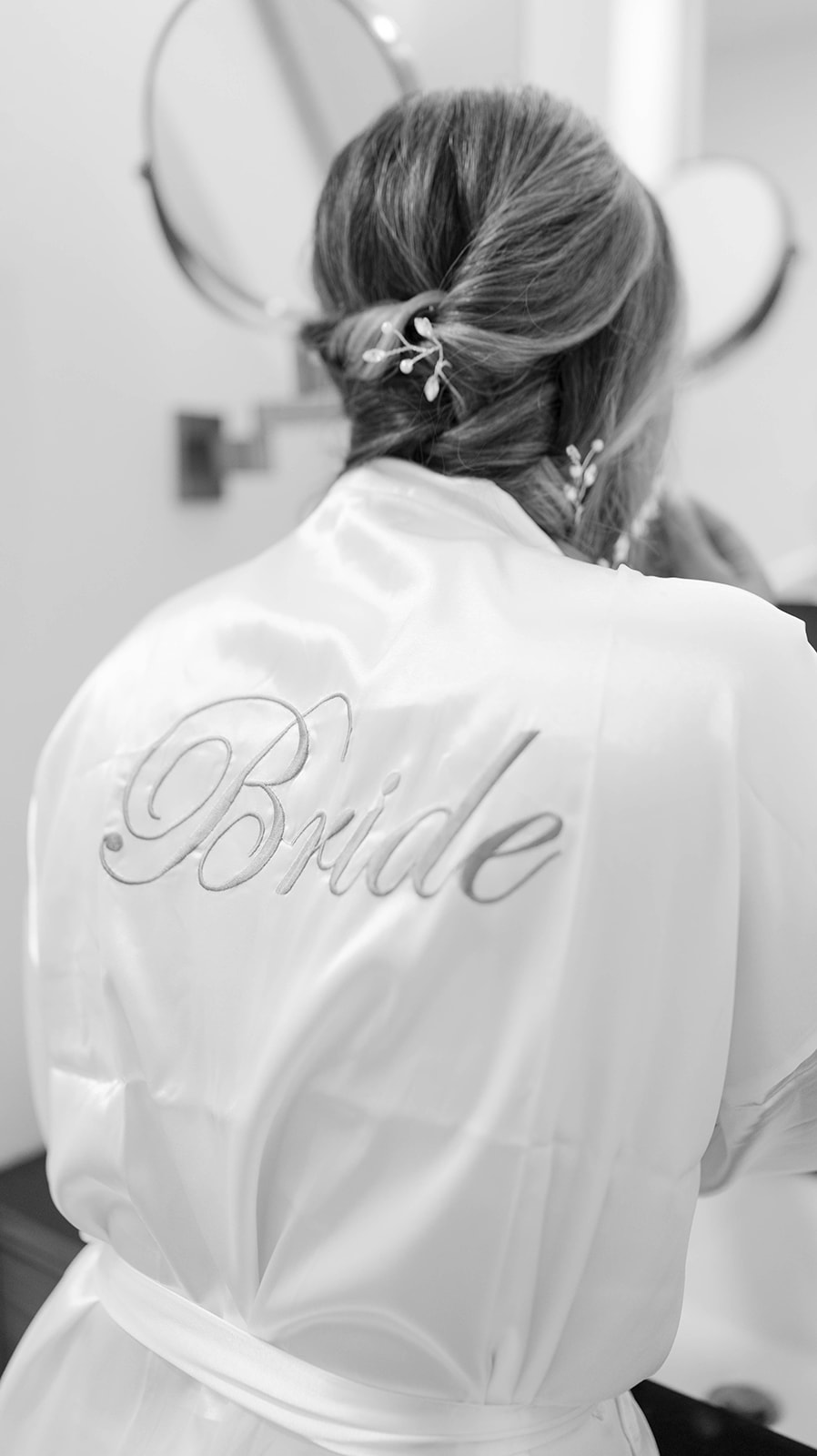 Black and white photo of bride getting ready, hair styled with delicate floral pins, robe embroidered with 'Bride.'