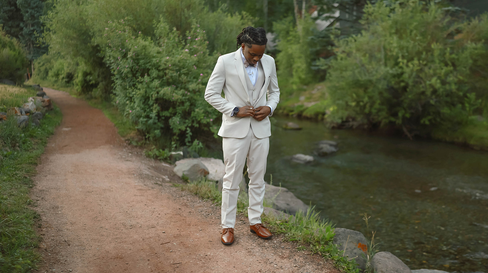 Groom in a cream suit buttoning his jacket on a dirt path by a creek, surrounded by lush greenery.