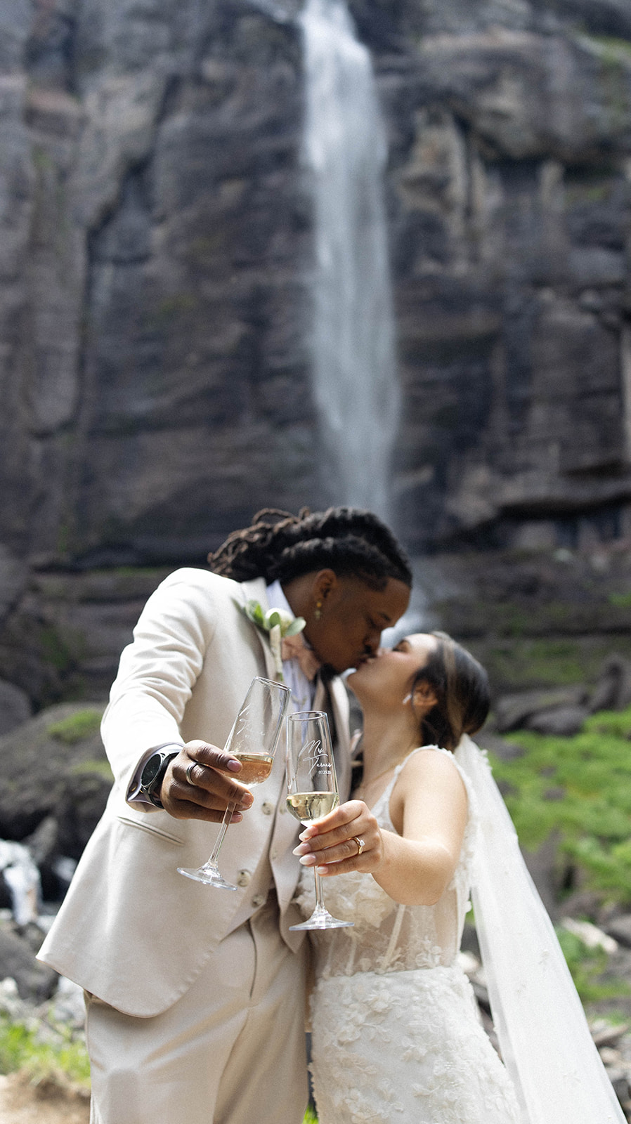 Couple clinking champagne glasses and kissing in front of a waterfall, celebrating their elopement.