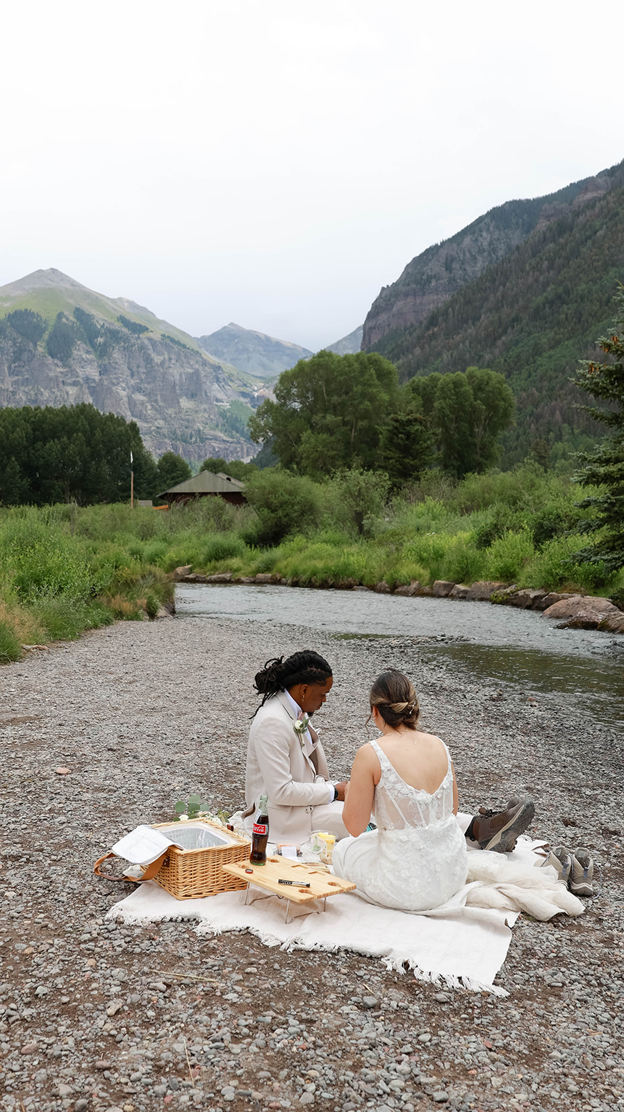 Bride and groom enjoying a picnic by the river, surrounded by mountains and greenery.