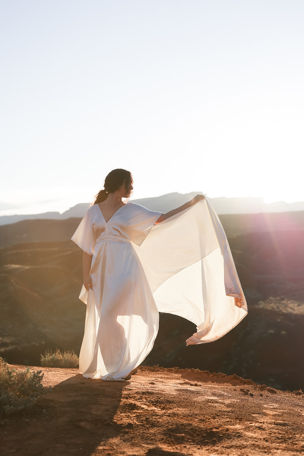 Bride standing on a cliff edge at sunset, holding her gown in the breeze, the desert landscape stretching out behind her in Castle Valley, Utah.