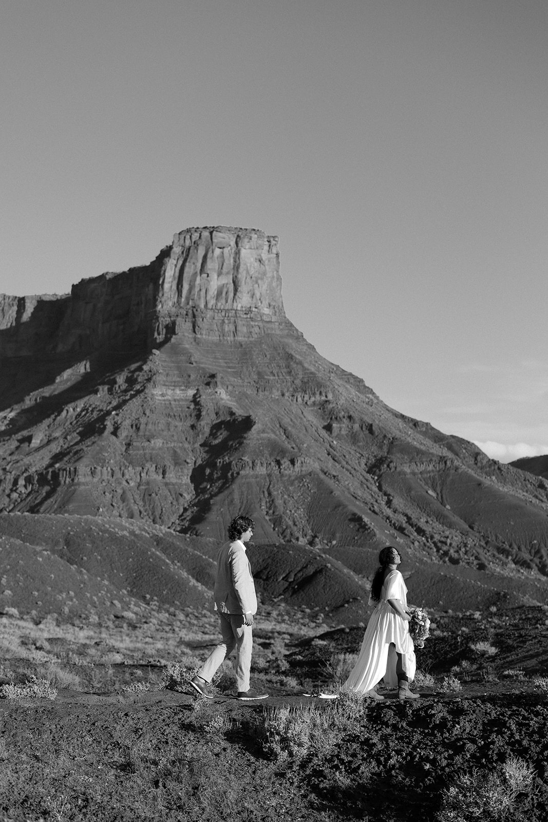 Black and white photo of a couple walking in front of a massive desert mesa, capturing the quiet beauty of elopements in Castle Valley, Utah.