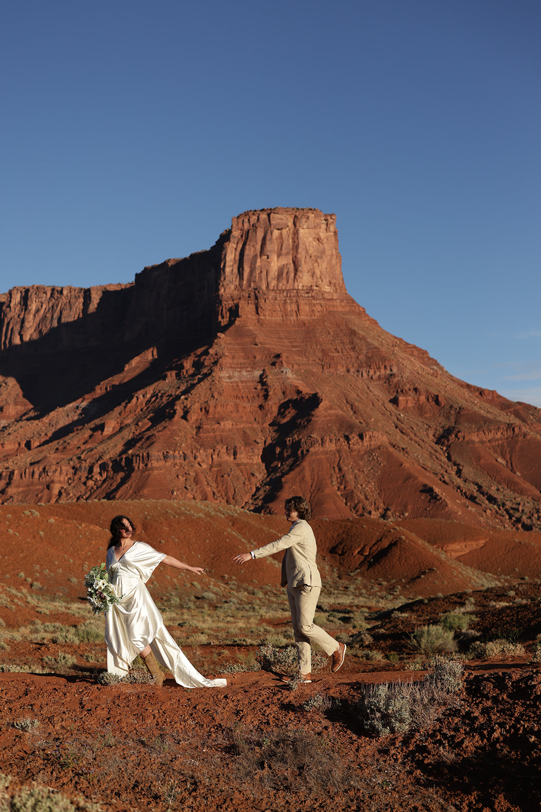 Bride playfully leads the groom across the desert with a dramatic mesa behind them, highlighting the adventurous spirit of Castle Valley, Utah.