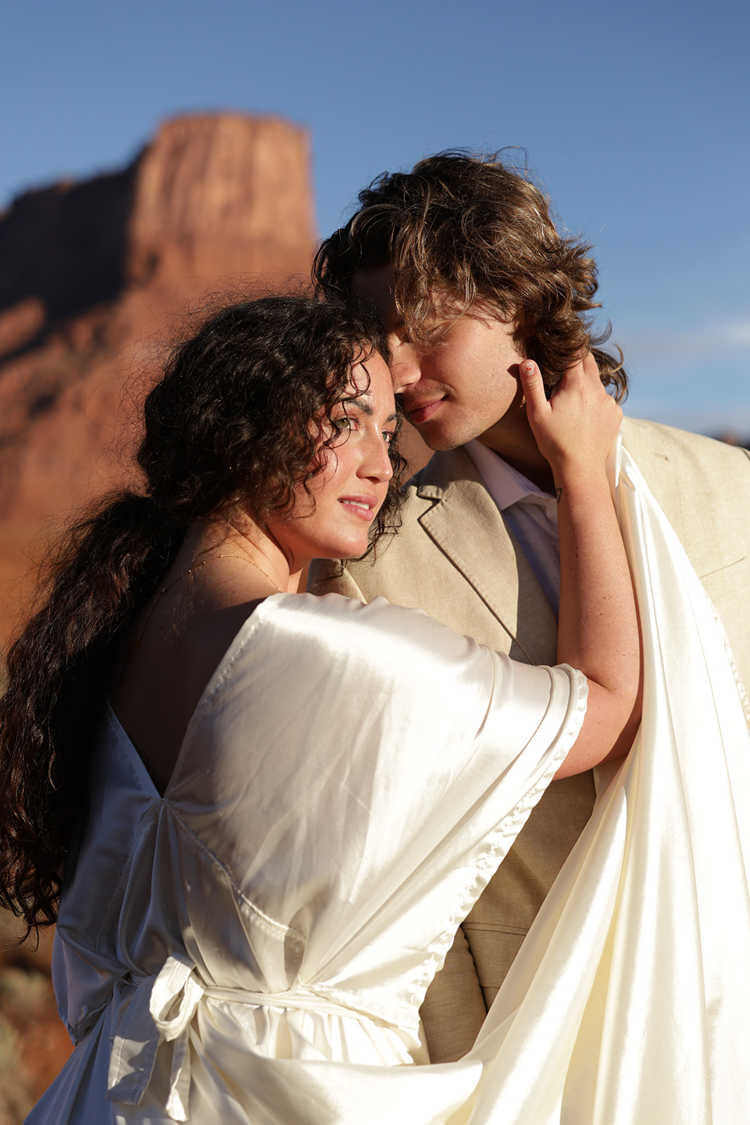 Tender close-up of a couple embracing in the desert, sandstone cliffs glowing red in the background.