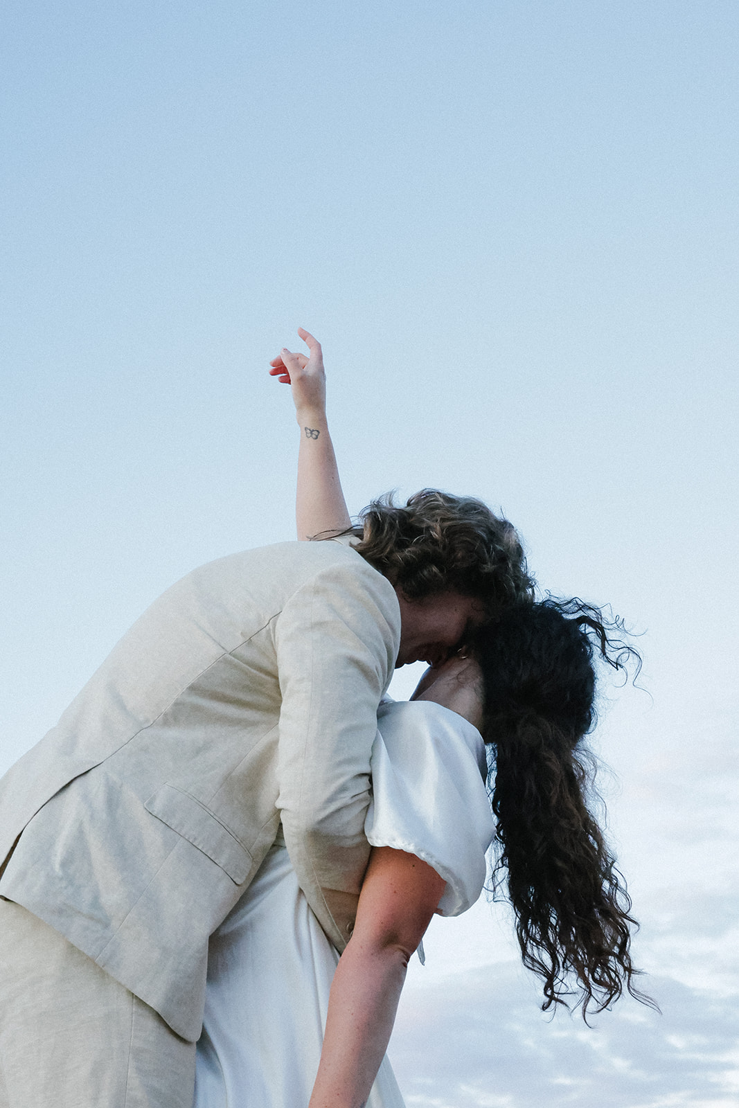 Close-up of a couple kissing at sunset, bride lifting her arm in celebration with blue sky overhead.