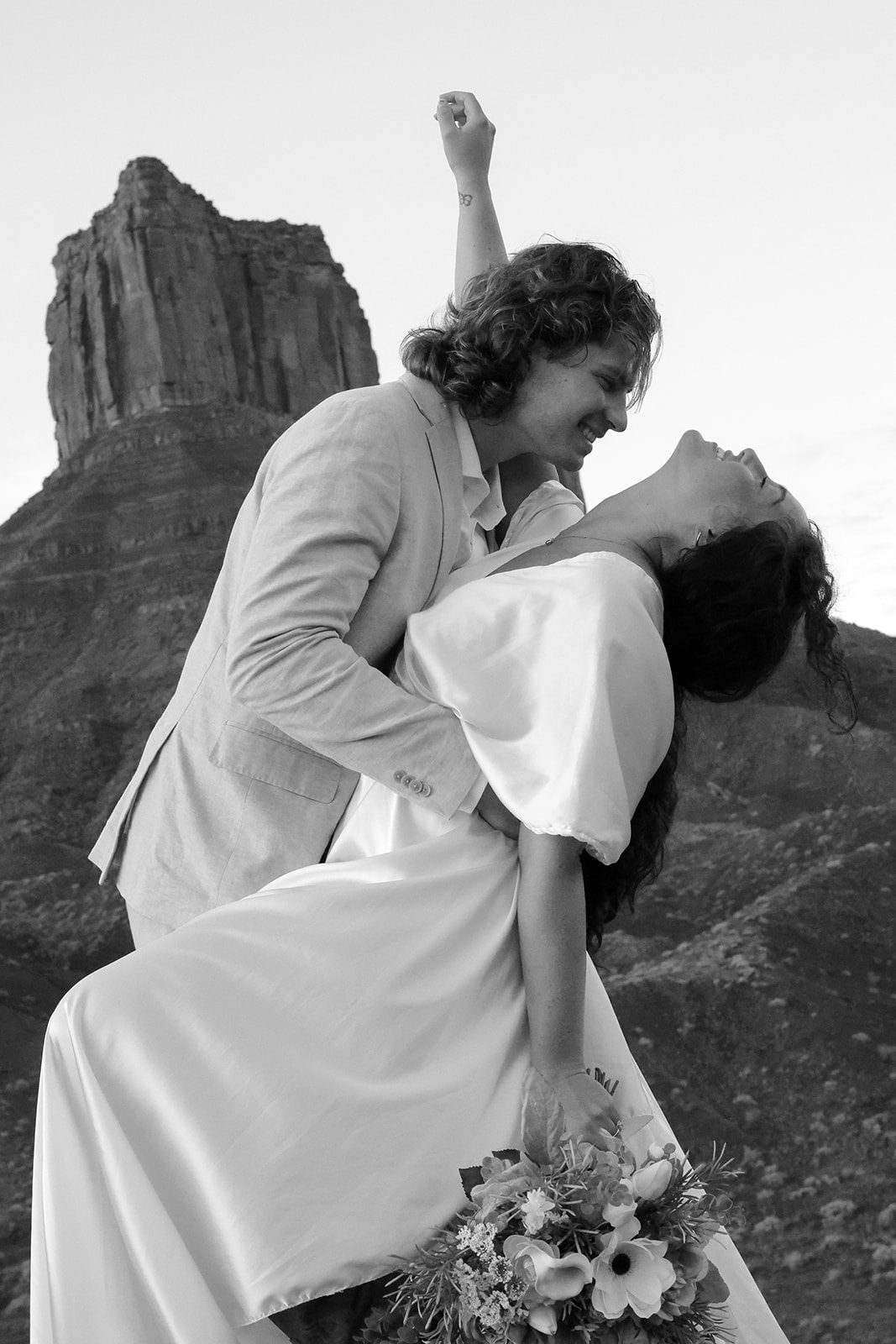Playful moment of a couple laughing and leaning into a dip with a towering mesa behind them in Castle Valley, Utah.