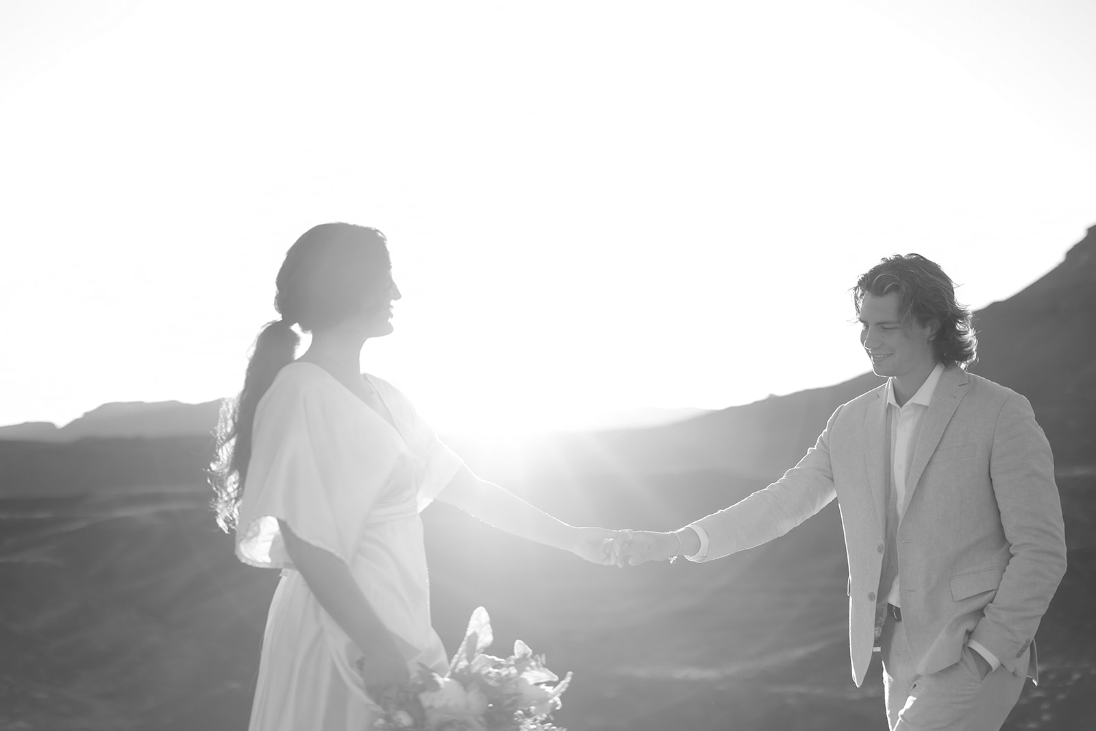 Black and white photo of a bride and groom holding hands with the sun shining behind them during their desert elopement.