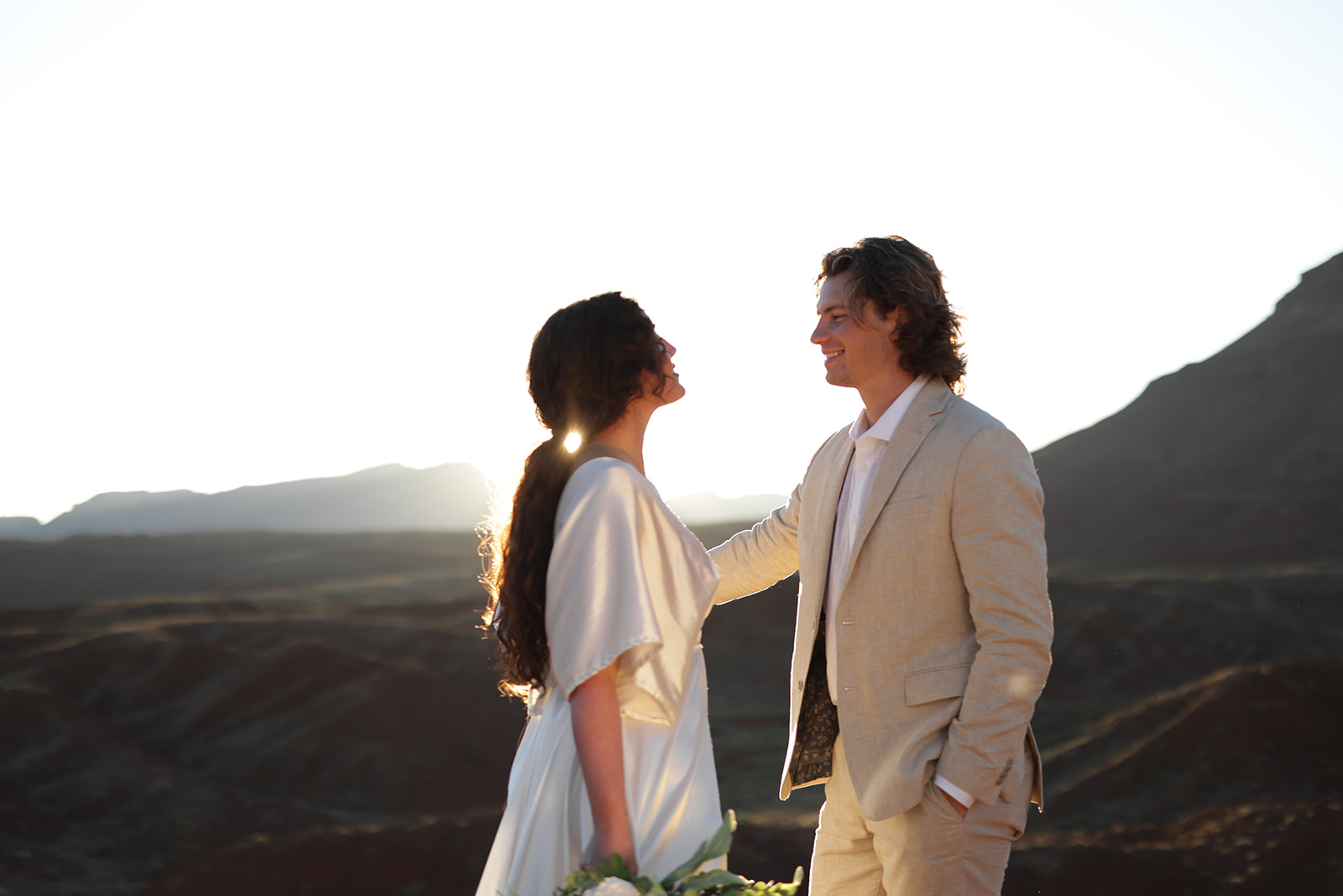 Bride and groom smiling at each other with the sun dipping behind the mountains, desert horizon glowing warmly.