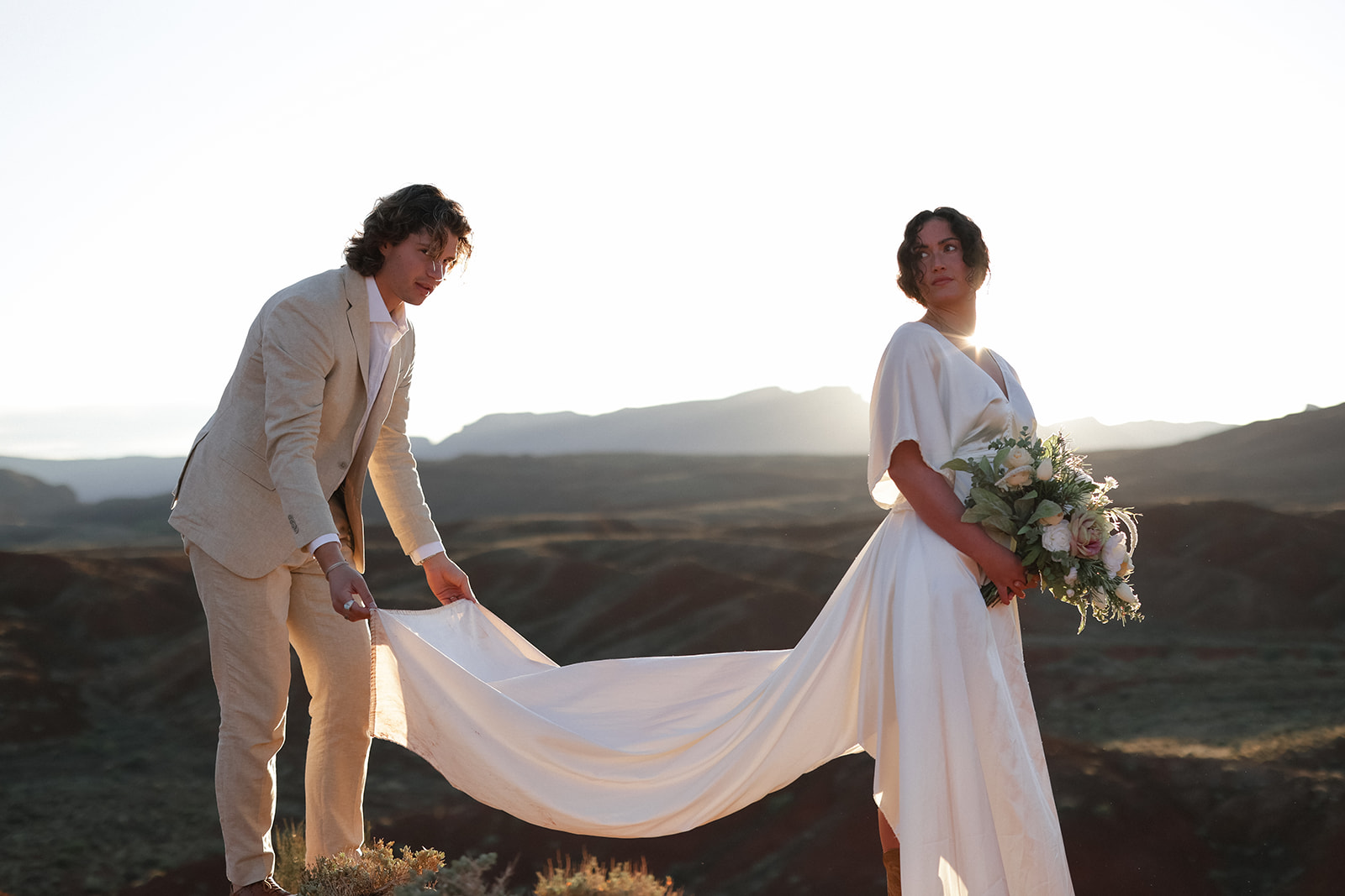 Groom holds the bride’s long flowing dress while she stands with bouquet, sunlight streaming behind them
