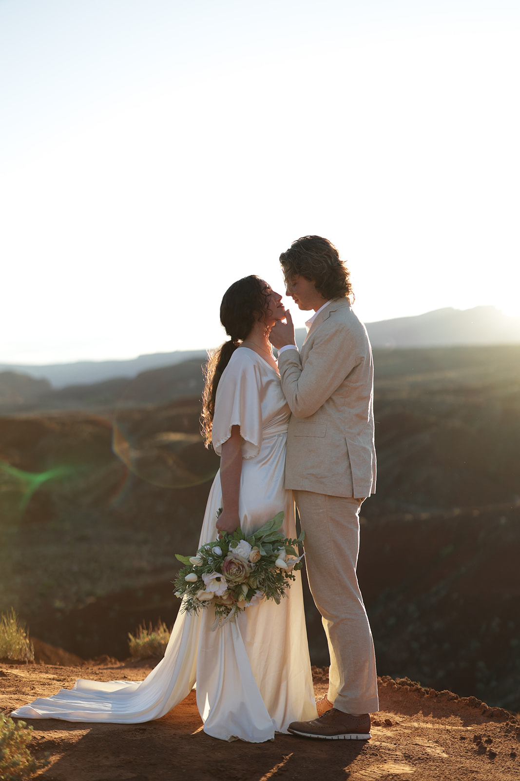 Couple standing face to face at golden hour on a cliffside, bride holding a lush bouquet while the groom touches her face gently.