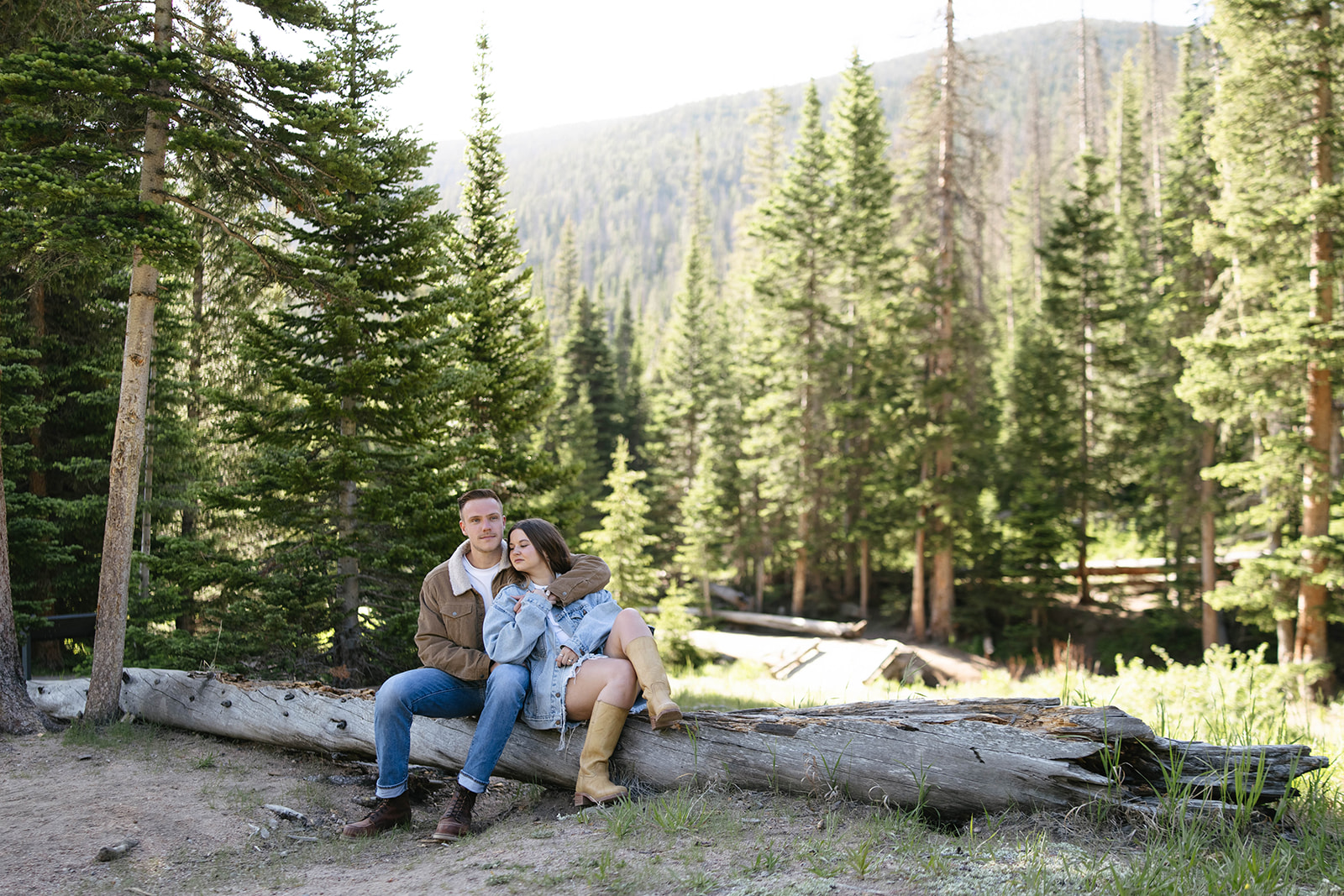 A couple sitting on a fallen log surrounded by tall pine trees, the mountains in the background creating a serene mountain wedding setting.