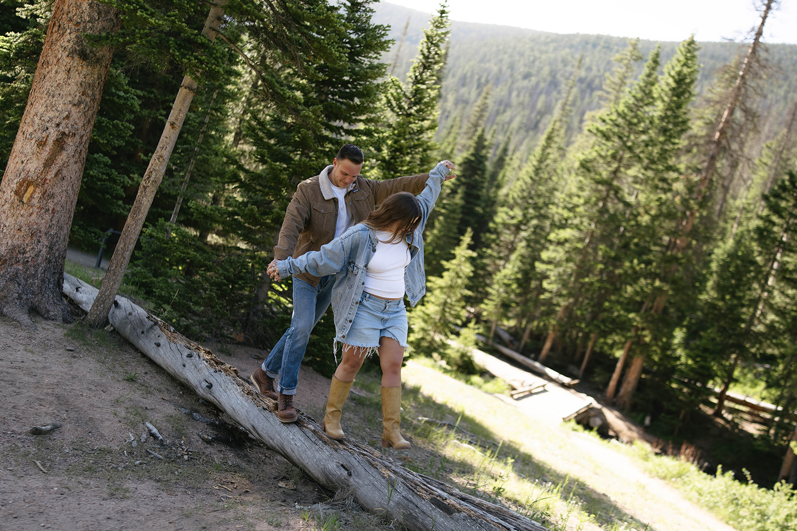 A joyful couple balancing on a log in the forest, holding hands with sunlight streaming through the pines.