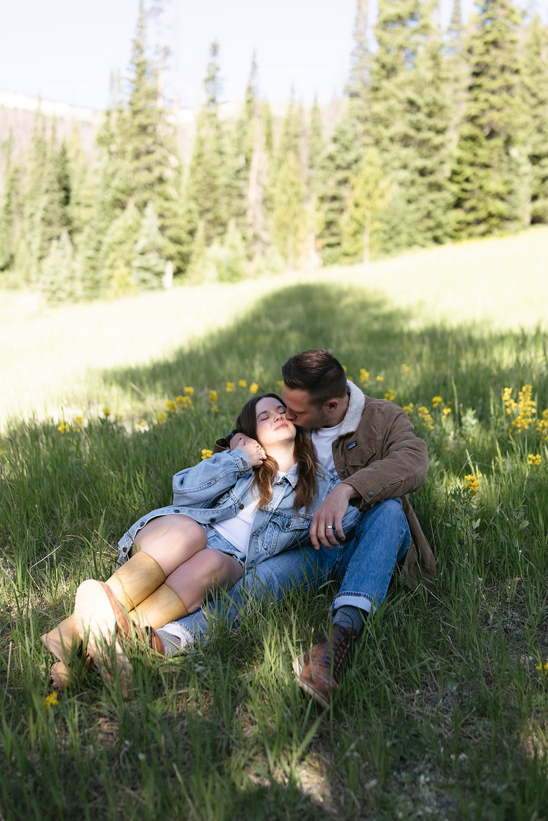 Man kisses his partner’s forehead while they relax in the grass, capturing a cozy mountain wedding–style vibe.
