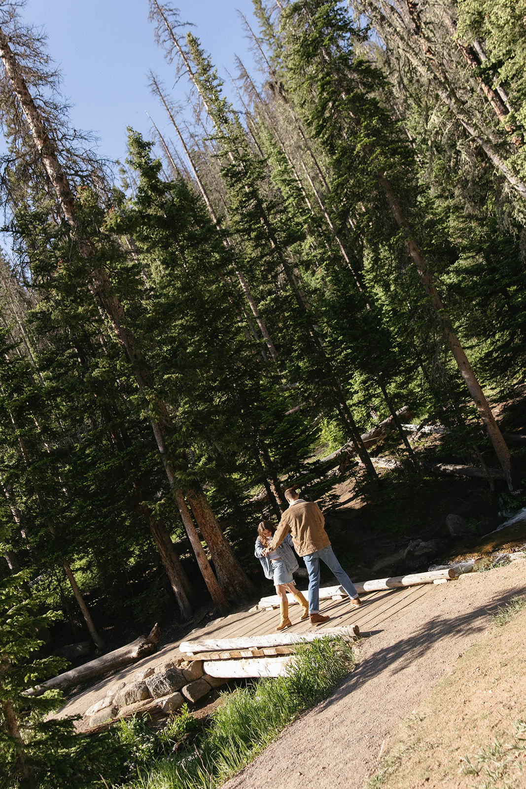 Playful moment of a couple dancing together on a wooden bridge in the forest, sunlight streaming through the trees.