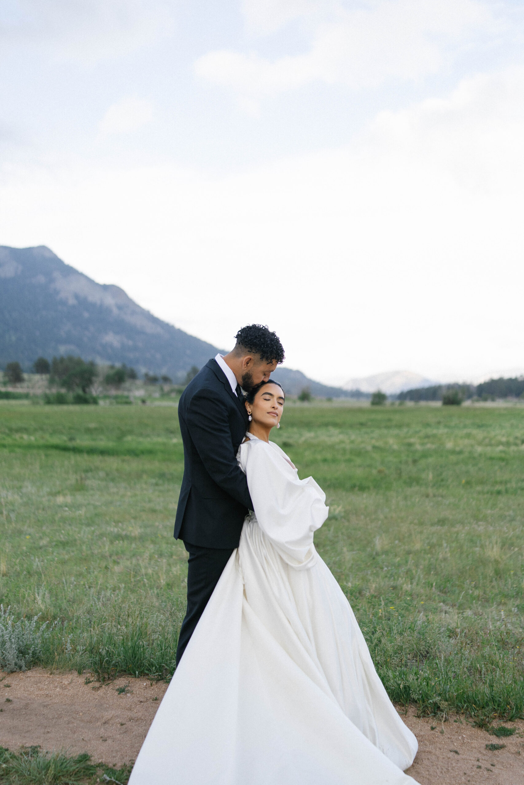 Groom holding his bride close in an open meadow with mountains in the background during their mountain wedding.