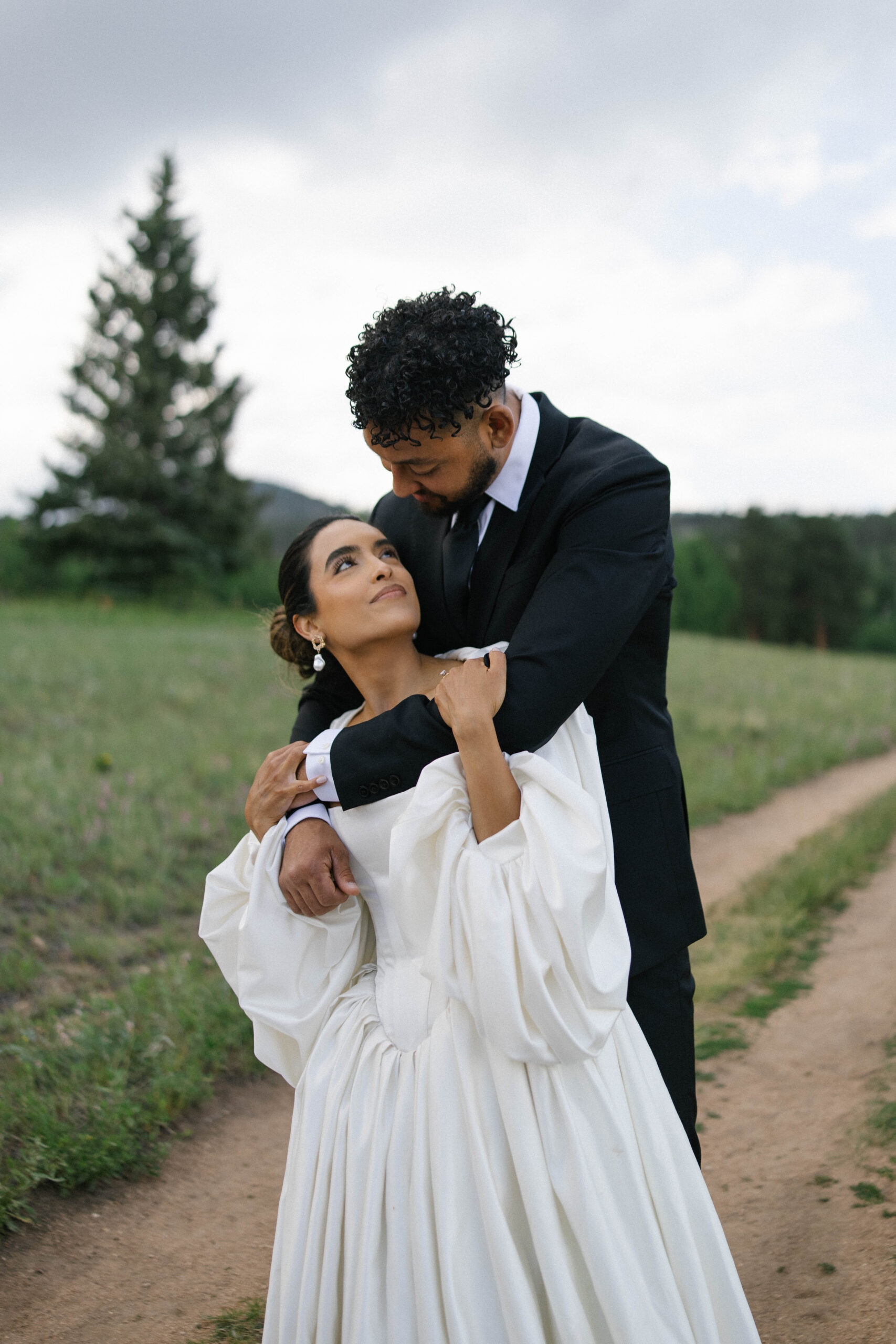 Bride looking up at her groom while he embraces her along a dirt path lined with greenery.