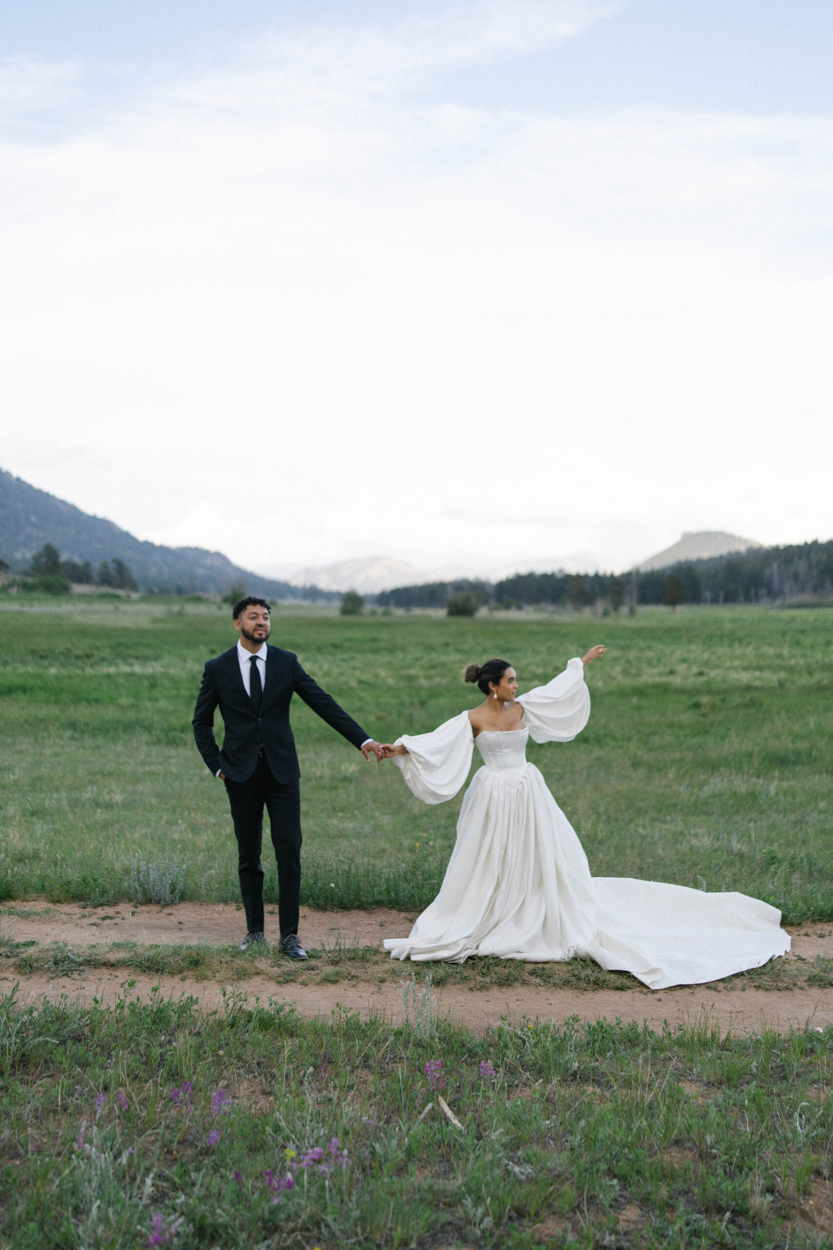 Couple holding hands and walking in a field, bride in a flowing gown with dramatic sleeves.