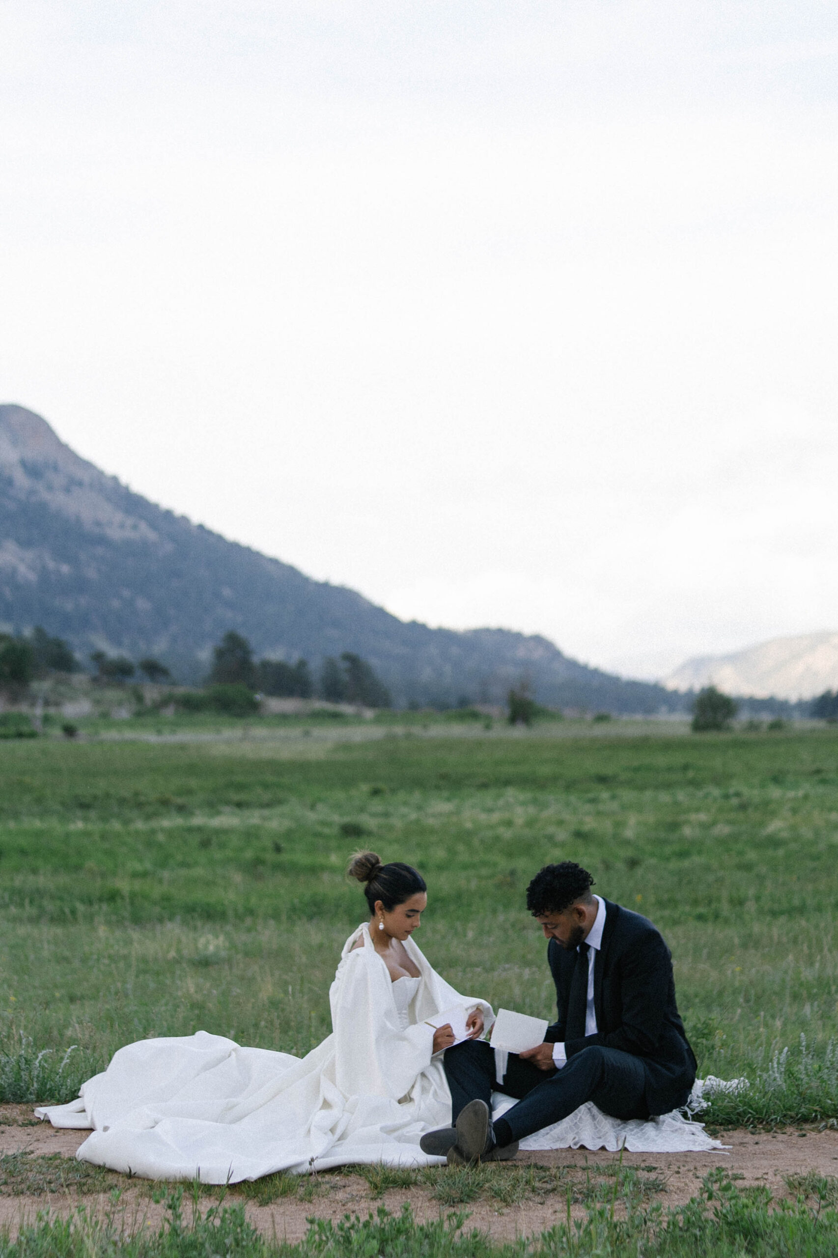 Bride and groom sitting together on the ground in an open field, reading vows to each other.