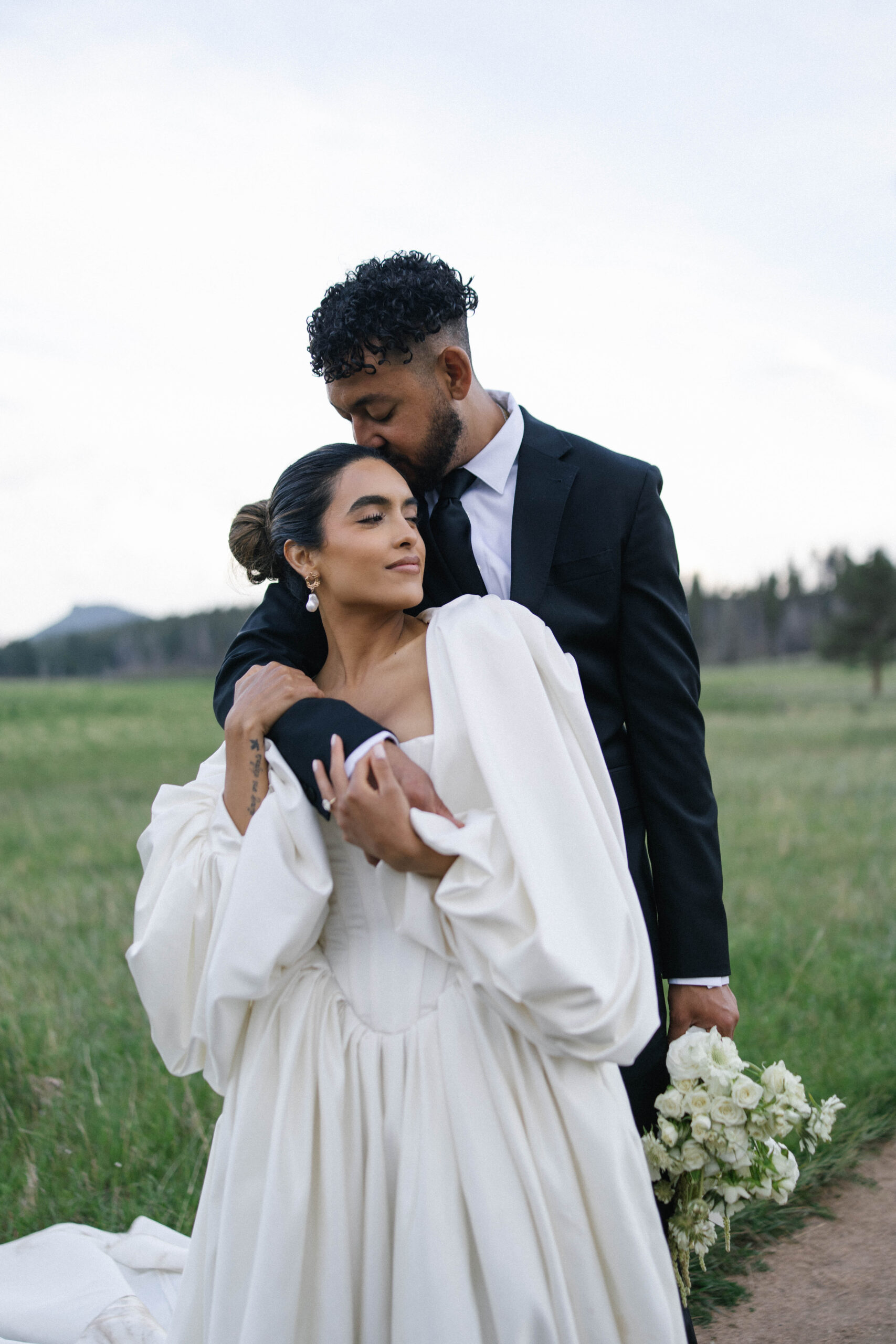 Groom kissing the bride’s head as she closes her eyes peacefully, holding her bouquet, a serene moment in their mountain wedding day.