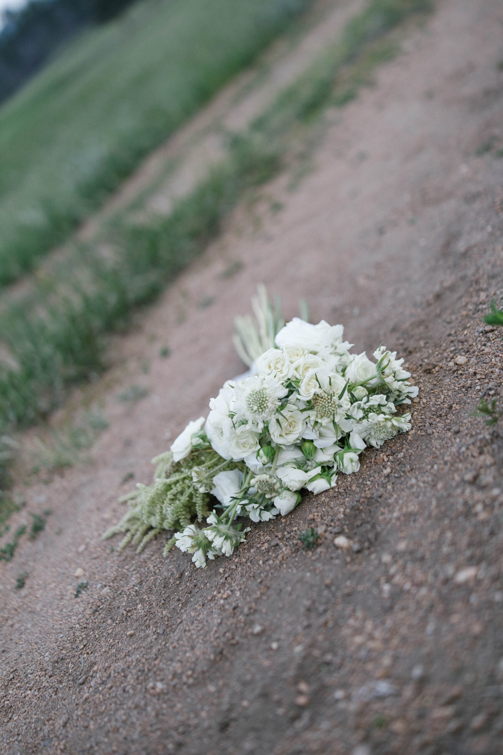 White wedding bouquet with roses and textured greenery resting on a dirt path.