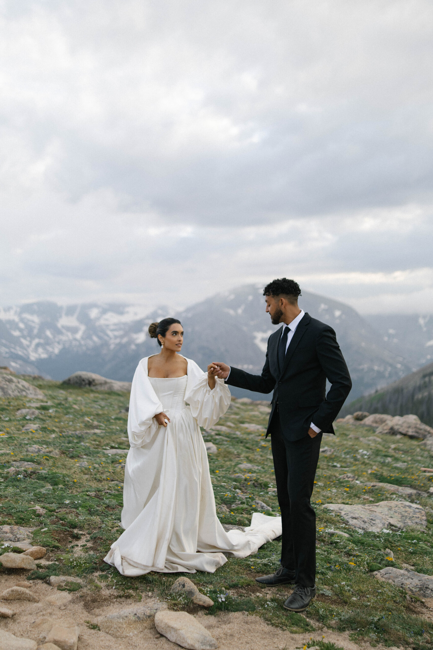 Groom holding the bride’s hand as they walk on rocky terrain with snowcapped peaks behind them during their mountain wedding.