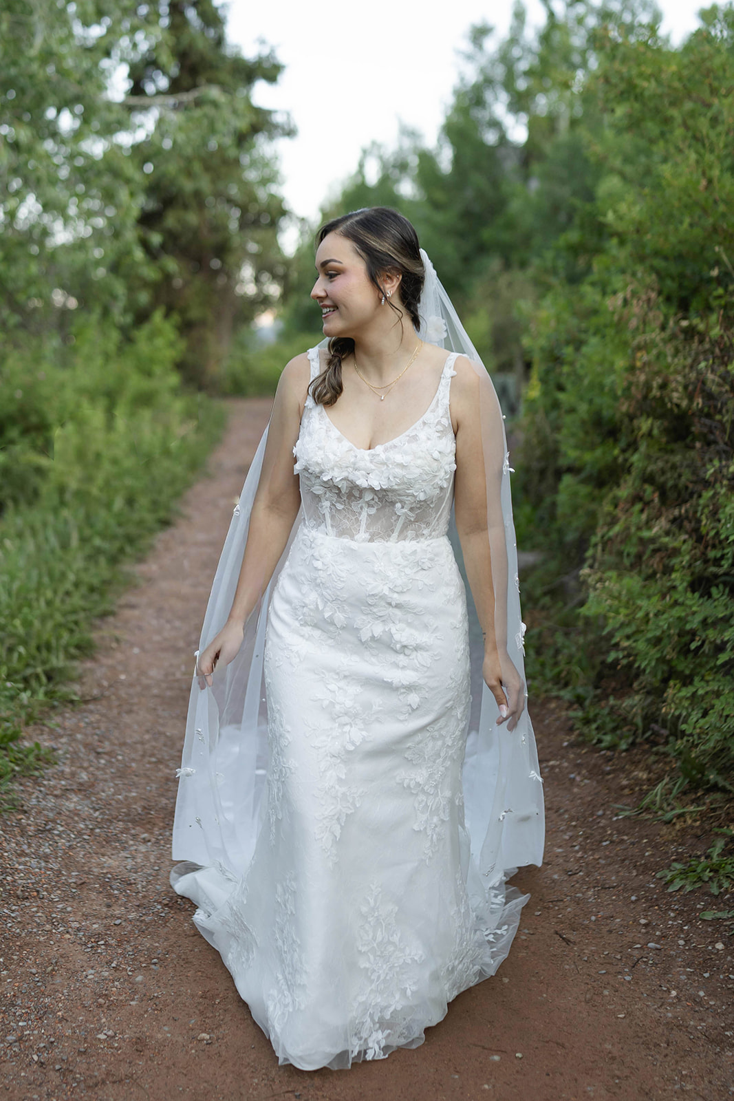 Bride smiles while walking down a dirt path in her lace gown and veil surrounded by greenery.