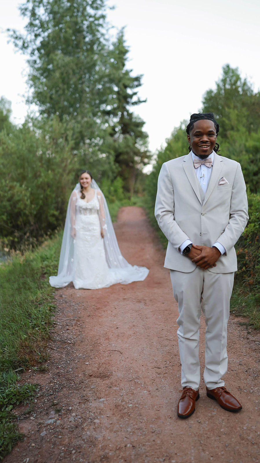Groom stands on a forest trail smiling while the bride in her gown approaches behind him, capturing a first look moment and sweet elopement ideas.