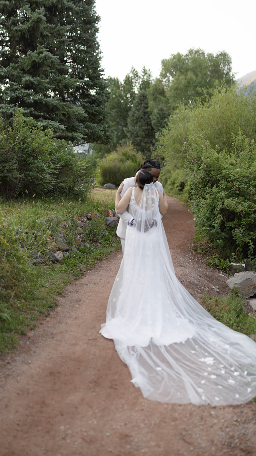 Bride and groom share an intimate hug on a forest path, her long veil flowing behind her.