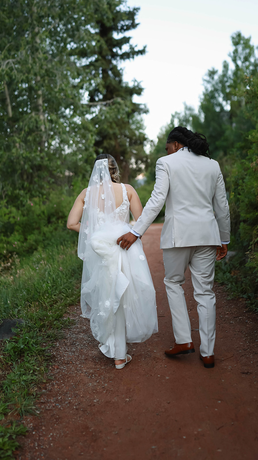 Bride and groom walk hand-in-hand down a wooded path, with the groom gently lifting the bride’s dress to keep it off the ground.