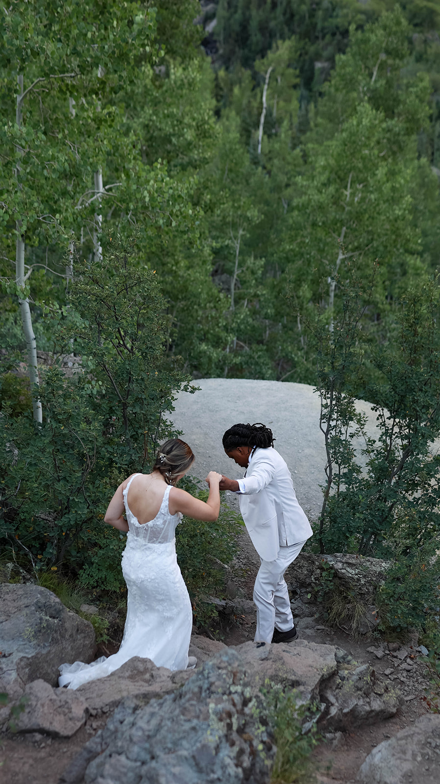 Bride in a lace gown being helped up rocky terrain by the groom in a cream suit during their mountain ceremony.