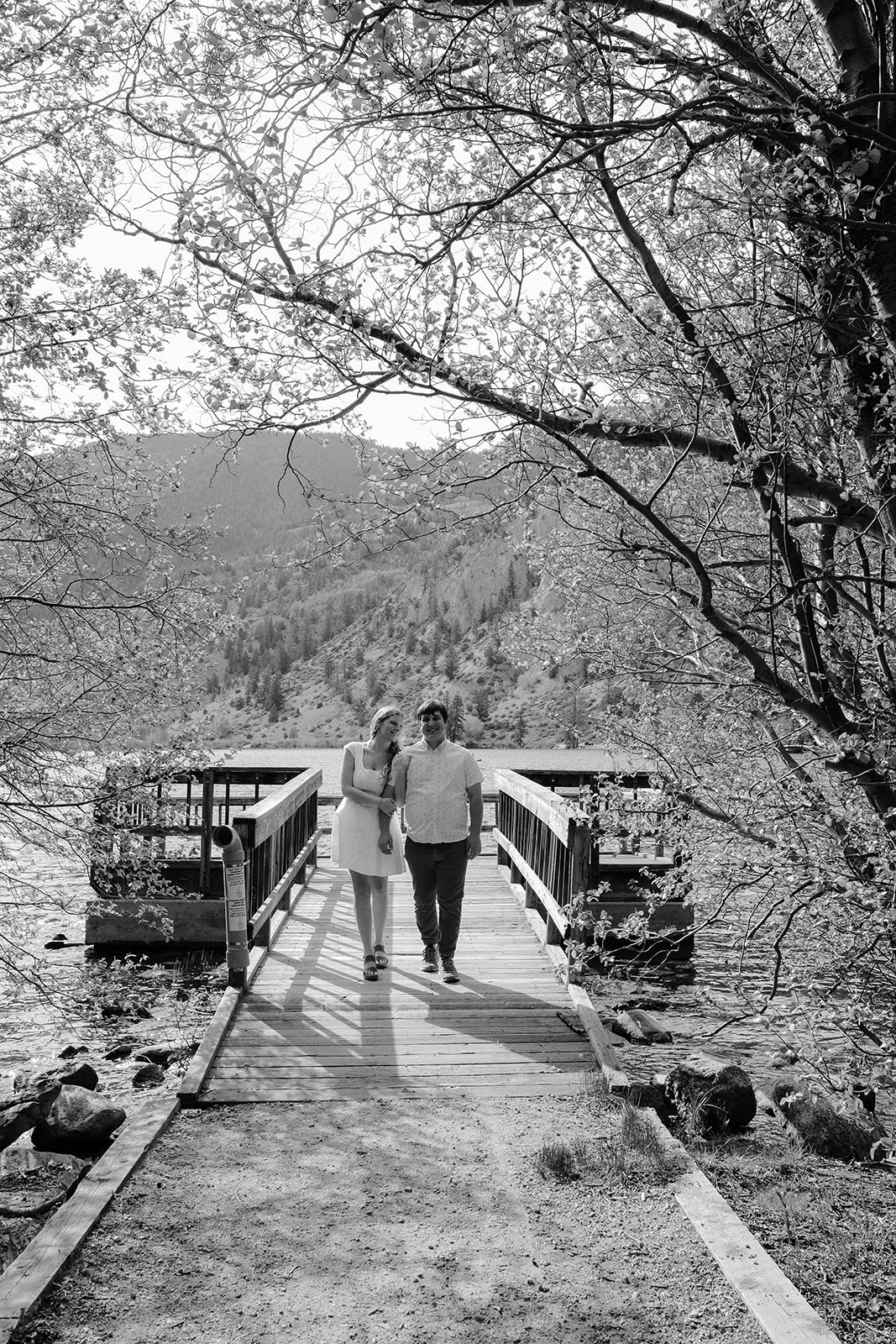 Black and white photo of a couple walking arm in arm down a dock with lake and mountains in the background in Buena Vista, Colorado.
