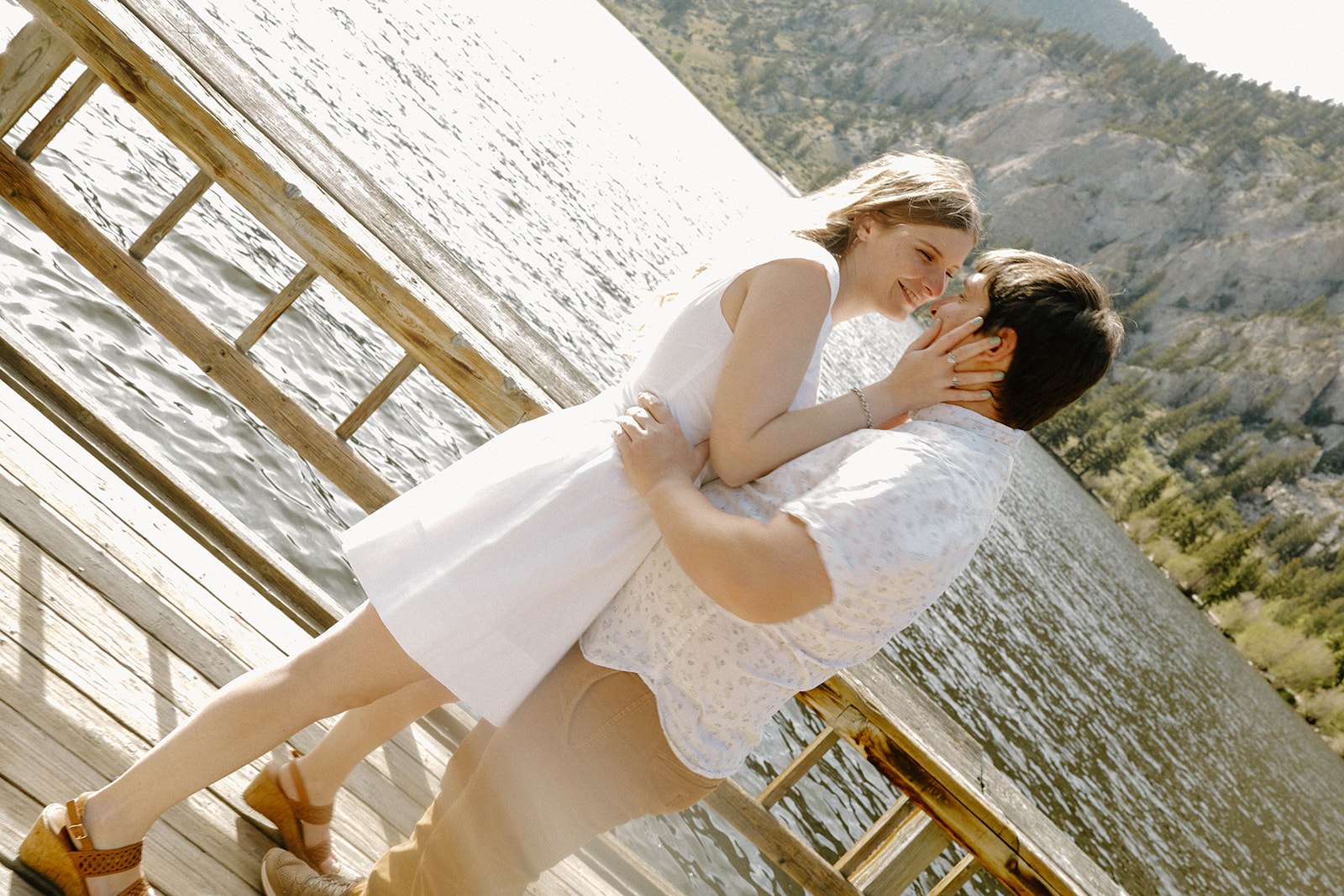 Romantic kiss on the dock as the couple leans into each other with the lake and mountains behind them.