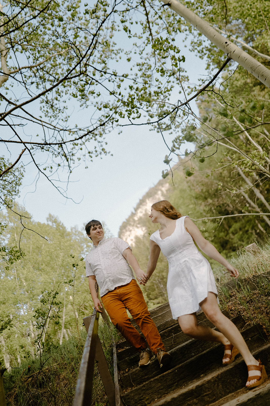 Couple holding hands and walking down rustic outdoor stairs surrounded by tall trees and sunlight.