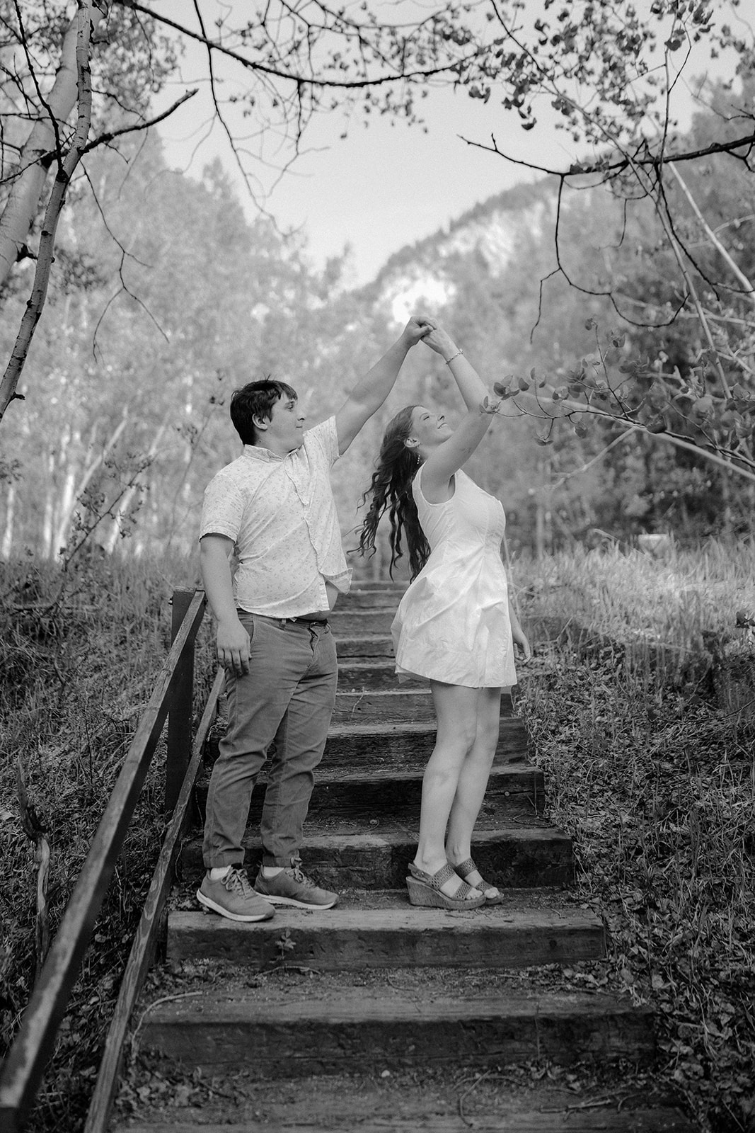 Couple twirling together on stone steps surrounded by trees, sharing a lighthearted moment.