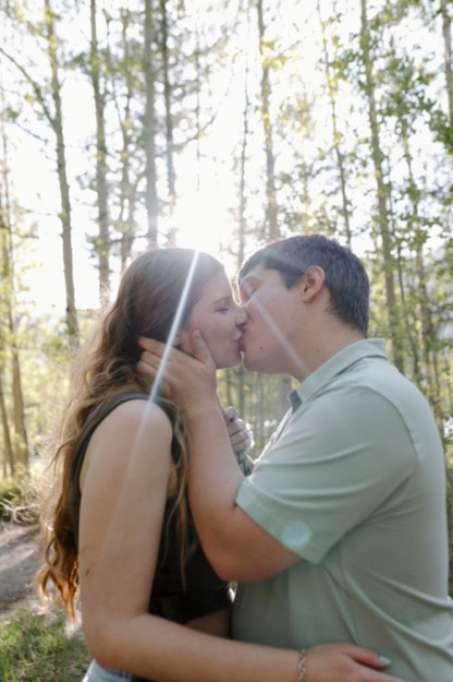 Couple sharing a kiss in the forest with golden sunlight streaming through the trees.