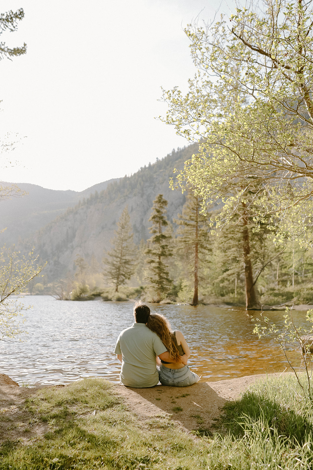 A couple sits on the grass at the edge of a lake, leaning into each other with mountains rising in the background.