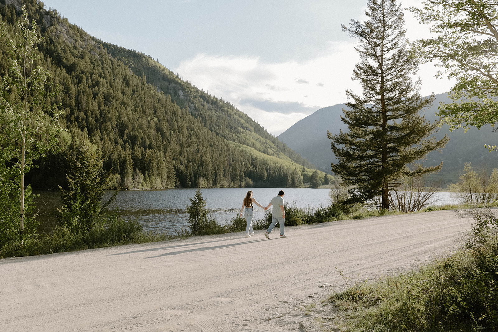 Couple walking hand in hand along a dirt road next to a mountain lake with tall pine trees and scenic views.