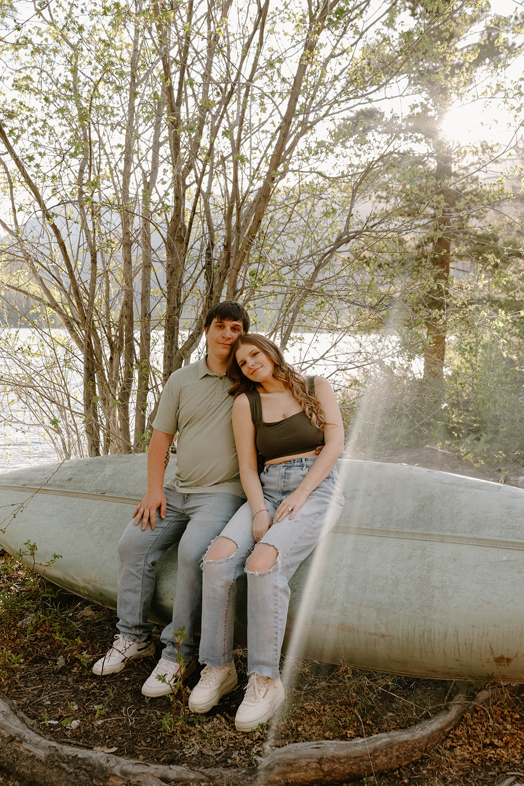 Romantic engagement photo of a couple sitting on a canoe, leaning close with the lake glistening in the background.