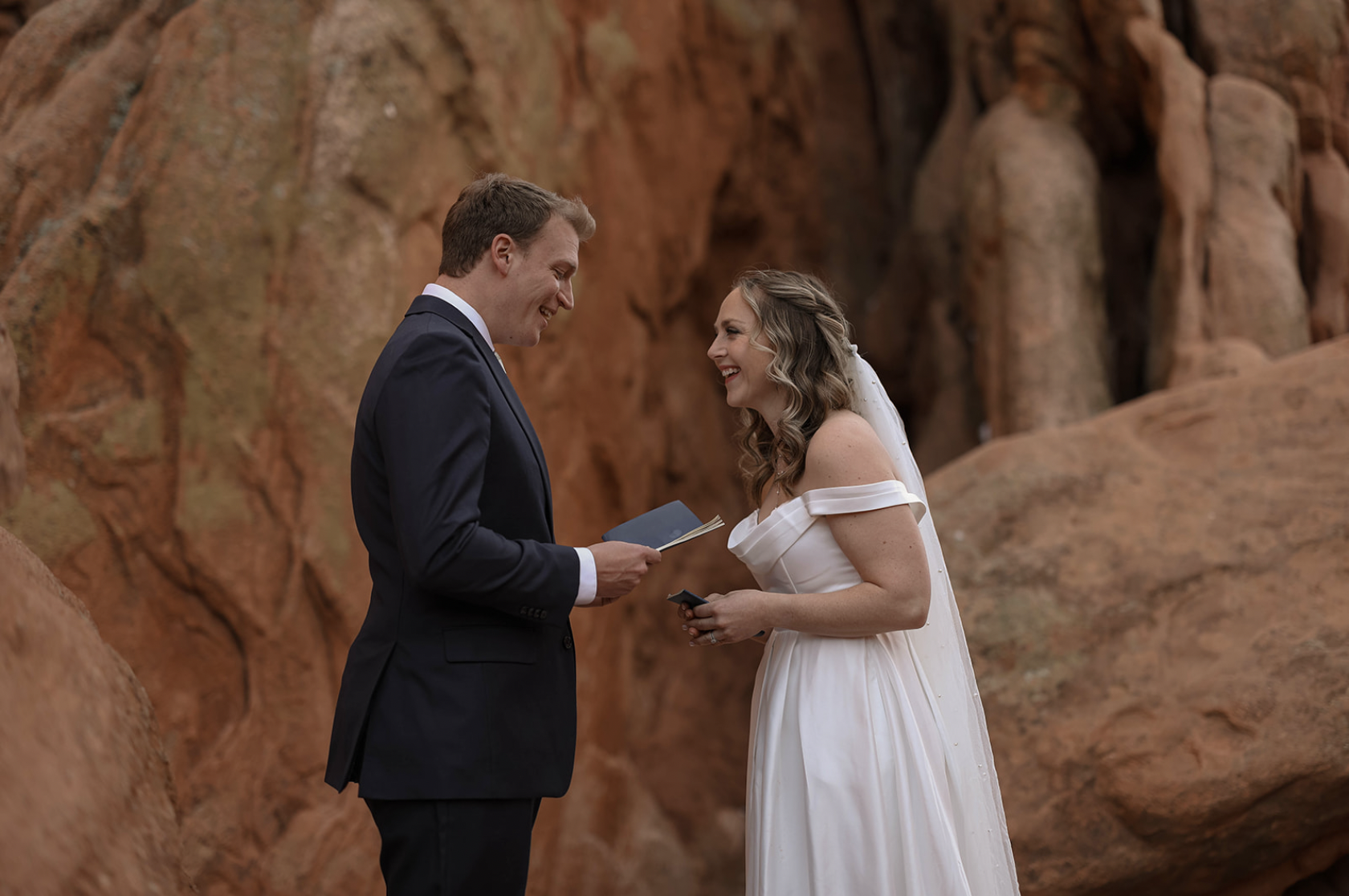A joyful bride and groom exchange vows in front of towering red rocks, smiling as they read to each other.