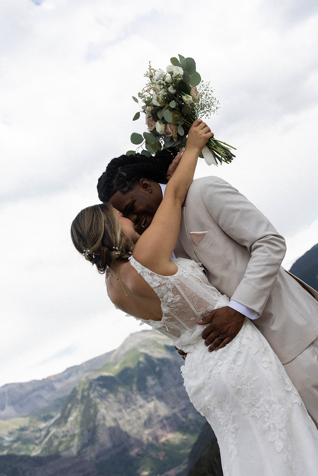 Bride and groom holding hands on a windswept alpine ridge, her voluminous white gown flowing around her while he stands in a sharp black suit. Colorado wedding photographer documenting adventure-filled moments.