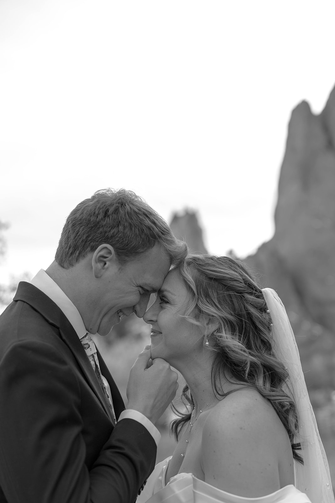 Black and white photo of a bride and groom touching foreheads and smiling, framed by rock formations in the background.