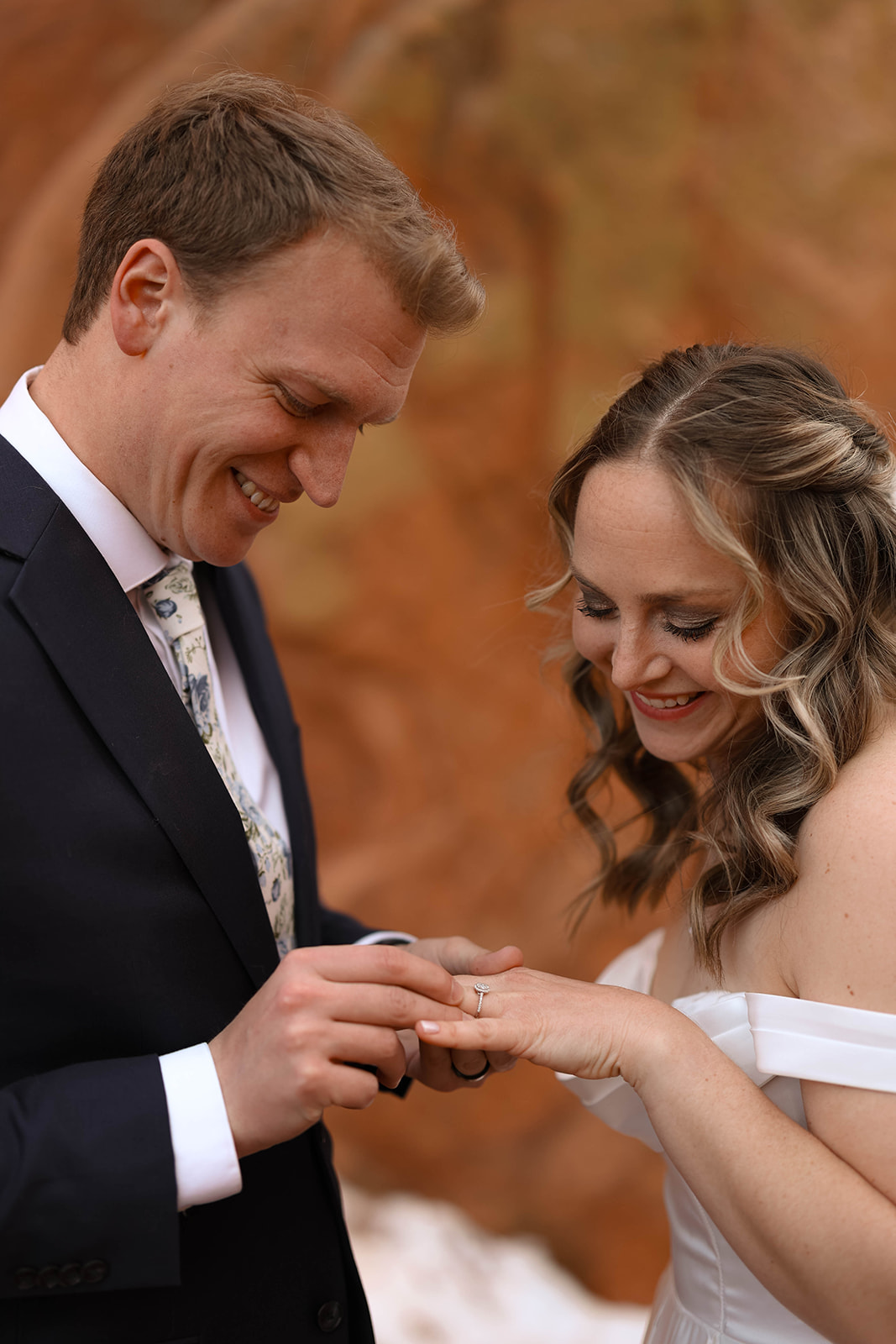 Bride and groom standing in front of towering red rock formations, the groom gently putting on his bride's wedding ring.