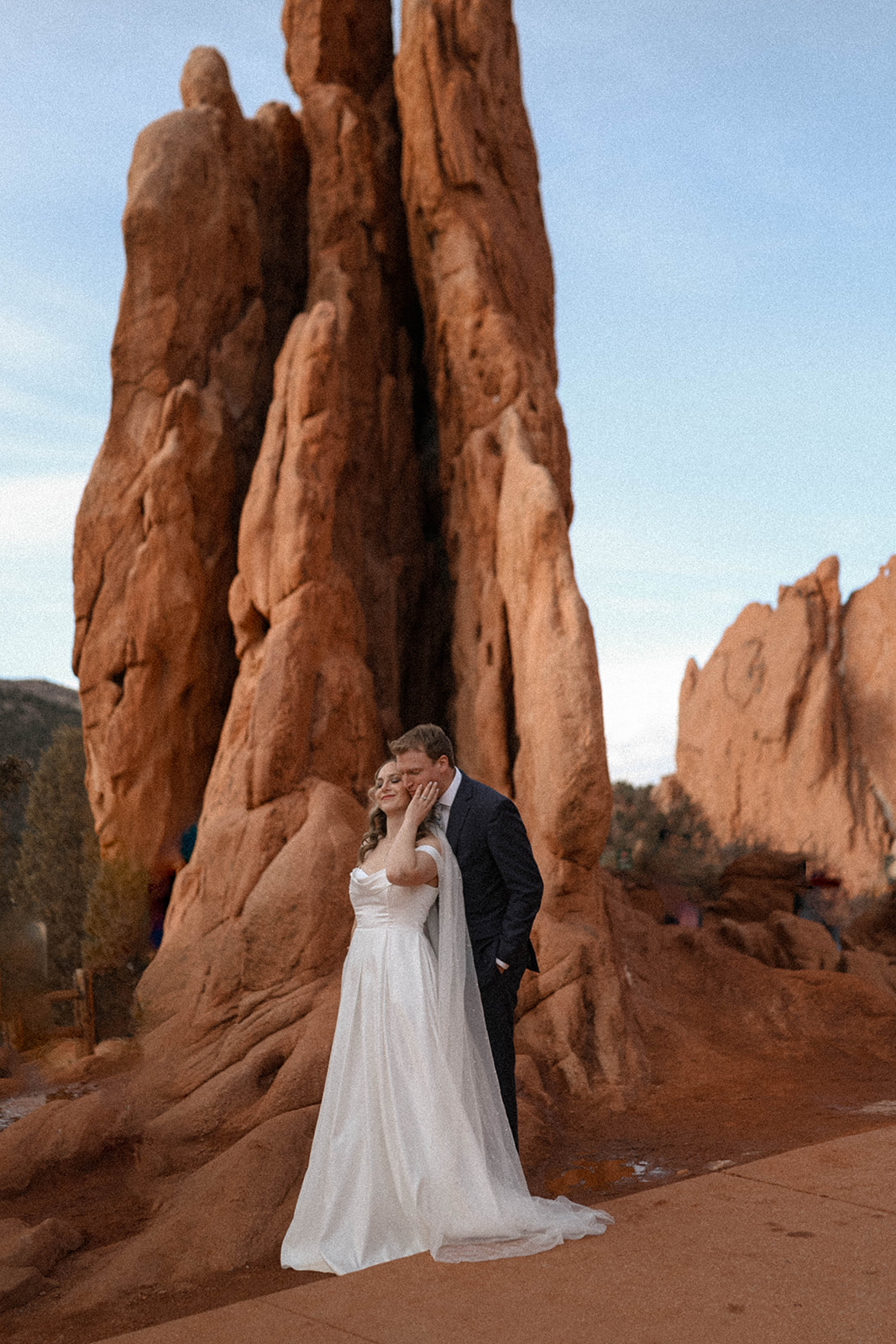 A bride and groom stand nestled against the red rock formations of Garden of the Gods, her flowing veil cascading while he kisses her temple.