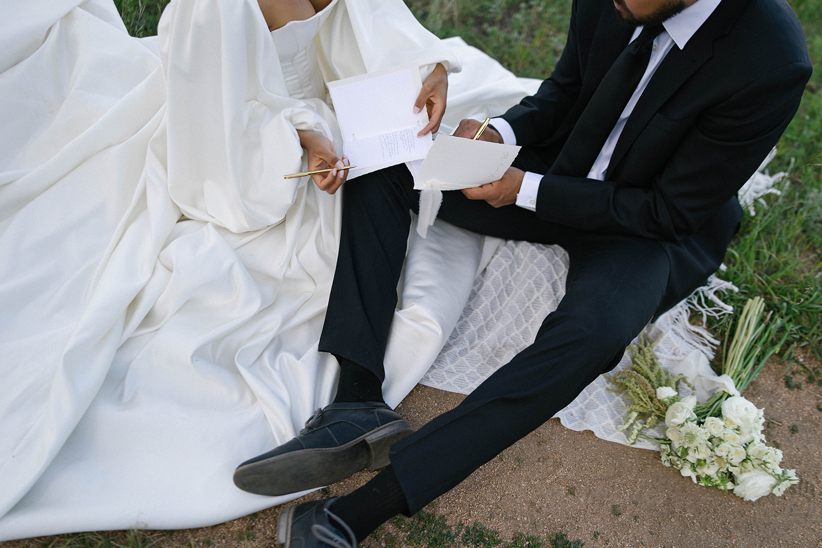 A couple sits close together on a blanket in the grass, writing their wedding vows by hand. Their outfits contrast beautifully with the natural setting, with the bride’s gown cascading across the ground.